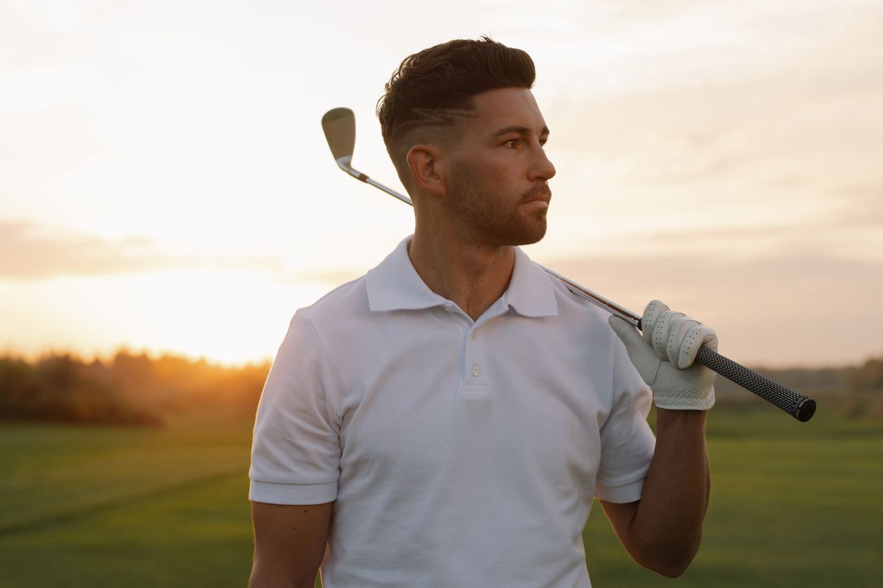 Portrait of a stylish male golfer posing with a club at sunset on a golf course, wearing a white polo shirt.