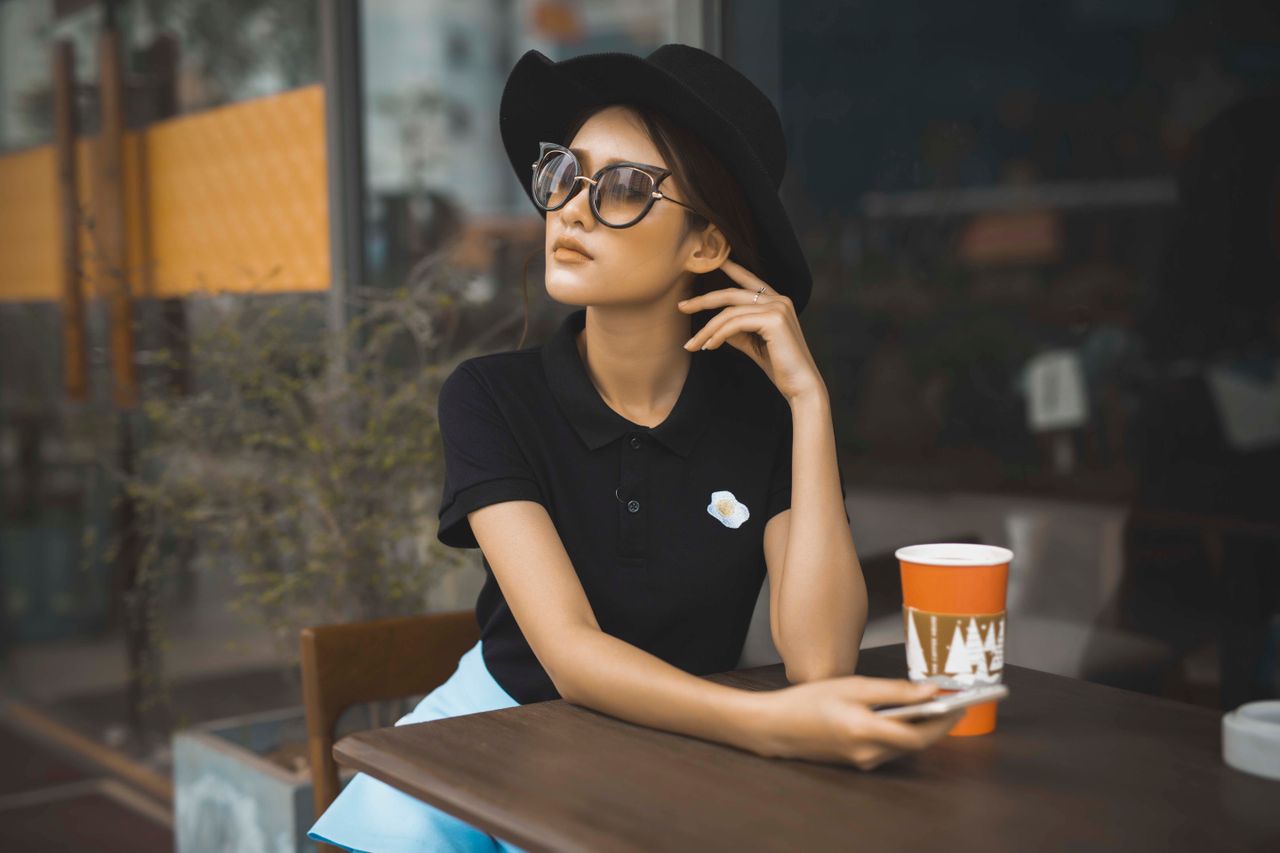 Chic woman in black hat and sunglasses sitting at a cafe with a coffee cup, embodying casual elegance.