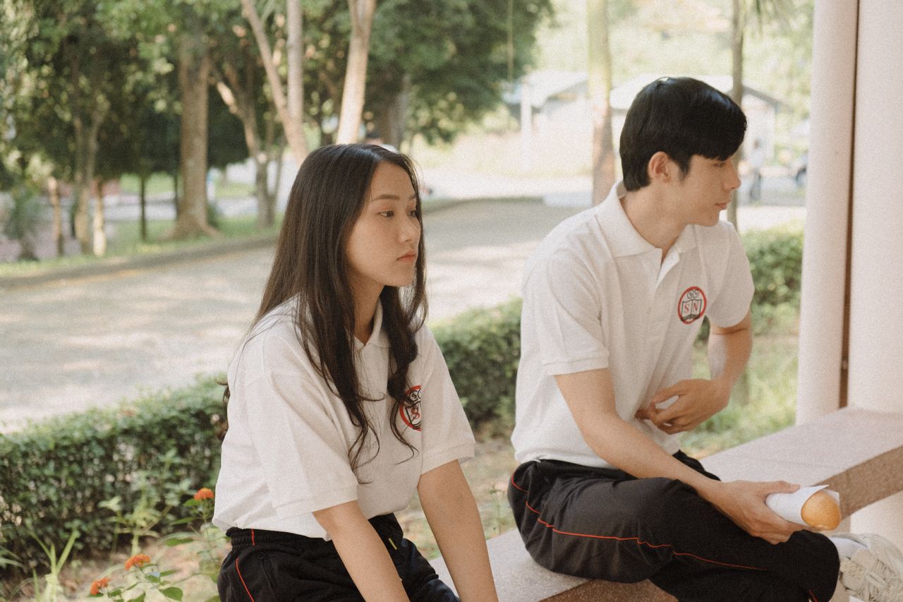 Two young adults sitting on a park bench in casual attire outdoors.